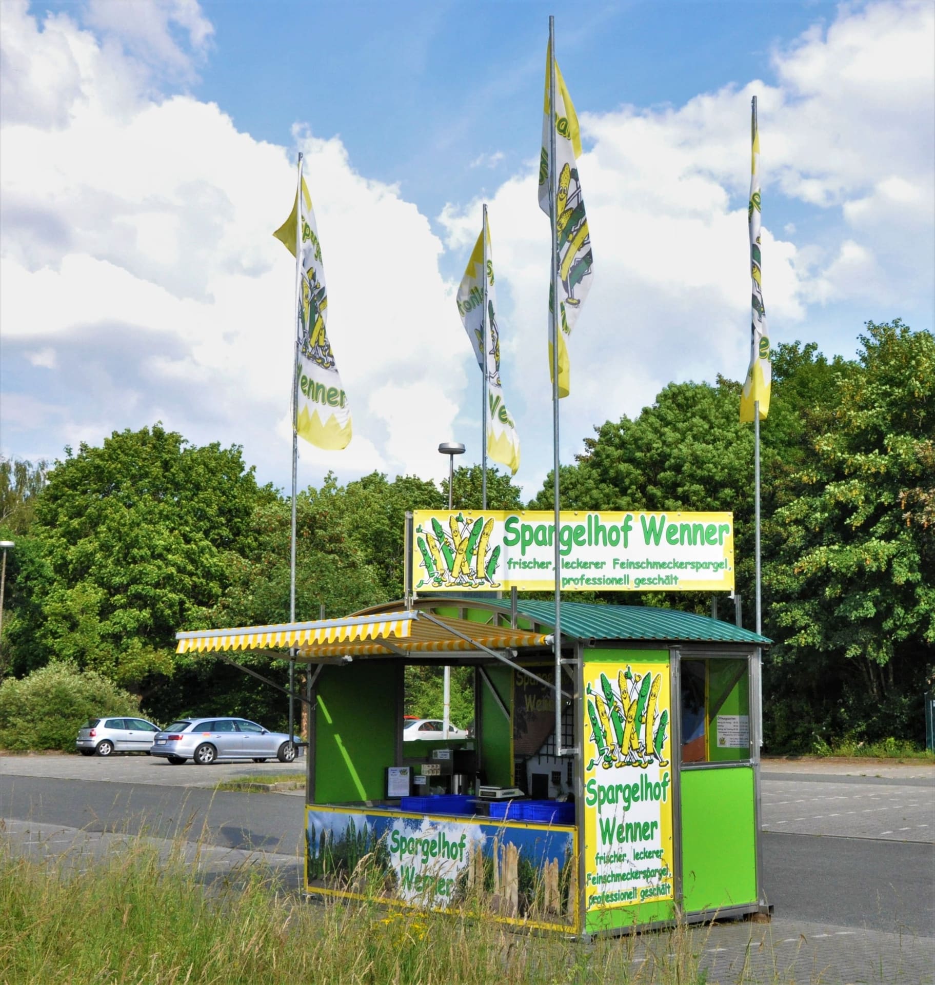 Ein Verkaufsstand im neuen Modell, grün mit gelb-weiß gestreifter Markise, Schild „Spargelhof Wenner“ und Fahnen